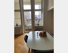 Innenraum eines modern eingerichteten Wohnbereichs mit großem Esstisch und Stühlen auf einem Parkettboden. Große Doppelfenster bieten Blick auf eine belebte Straße mit Gebäuden; draußen ist ein kleiner Balkon sichtbar.