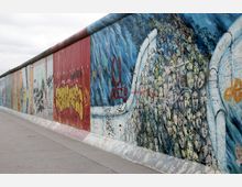 Ein Abschnitt der Berliner Mauer, bemalt mit bunten Graffiti und Wandmalereien. Die Motive zeigen unter anderem dicht gedrängte Gesichter und abstrakte Muster; der Boden besteht aus grauem Asphalt.