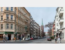 Straßenszene in einer Stadt mit historischen Gebäuden, Balkonen und Geschäften im Erdgeschoss; im Hintergrund ist eine Kirche mit einem spitzen, grünen Turm sichtbar. Autos parken entlang der Straße, und Passanten sind auf den Gehwegen unterwegs.