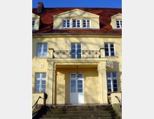 Gelbes Gebäude mit rotem Ziegeldach und symmetrischer Fassade, charakterisiert durch weiße Sprossenfenster und einen zentralen Eingangsbereich mit Säulen und einer gläsernen Laterne. Vor dem Eingang führen Steinstufen hinauf, und ein schmiedeeisernes Geländer ist über dem Portal angebracht.