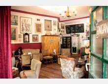 Gemütliches Café mit antiken Möbeln, Holzfußboden und einer gelben Kachelheizung. An den Wänden hängen zahlreiche Kunstwerke und ein Menü an einer Kreidetafel; im Hintergrund stehen Kühlgeräte und eine Theke.