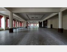 Weitläufiger, leerer Innenraum eines Gebäudes mit einer Reihe von großen Fenstern an der linken Seite, die rote Vorhänge haben. Die Decke ist flach mit eingelassenen Paneelen, und weiße Säulen unterteilen den Raum, während der Boden aus dunklem Fliesenmaterial besteht.