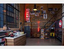 Industriehalle mit gelben Backsteinwänden, einem großen Stahlrahmenfenster und einer Metalltreppe. Neben Regalen mit Flaschen und Produkten befindet sich eine moderne Verkaufstheke aus Holz, während die Begriffe "Casino" und ein rotes Neonlogo die Wand dekorieren.