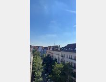 Straßenszene in einer Wohngegend mit Bäumen gesäumten Gehwegen, Altbauwohnungen mit dunklen Schrägdächern und einem Blick auf den Fernsehturm im Hintergrund unter klarem, blauem Himmel.