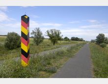 Ein gepflasterter Weg führt durch eine ländliche Wiesenlandschaft mit vereinzelten Bäumen und Wildblumen. Im Vordergrund steht ein schwarz-rot-gelb gestreifter Grenzpfosten mit der Nummer 522.