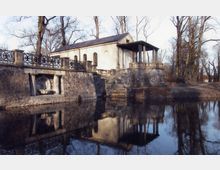 Historisches Gebäude im klassizistischen Stil mit Säulenveranda, steht an einem Teich mit Spiegelung im Wasser; daneben eine steinerne Ufermauer mit Ziergeländer und einer eingelassenen Wandnische mit Brunnen.