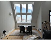 Moderner Essbereich in einem Dachgeschossraum mit schrägen Wänden und großem, mehrteiligem Dachfenster mit Blick auf die Stadt. In der Mitte stehen ein runder Tisch mit vier gepolsterten Stühlen auf einem Teppich, umgeben von hellem Holzboden und minimalistischer Beleuchtung.