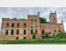 Eine verfallene Backsteinruine eines historischen Gebäudes mit einem rechteckigen Grundriss und einem angrenzenden Turm. Fensteröffnungen ohne Glas und teilweise überwachsene Vegetation im Vordergrund, mit Bäumen im Hintergrund.