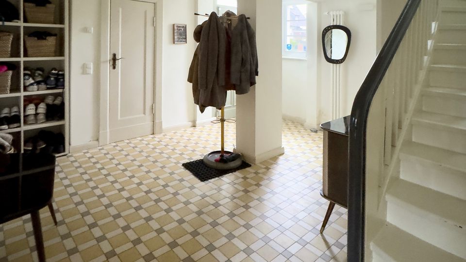 Heller Hausflur mit gemustertem Fliesenboden, weiß gestrichenen Wänden und einer Treppe mit dunklem Handlauf rechts. Links stehen ein offenes Schuhregal und eine Tür, in der Mitte eine Garderobe, hinten ein Fenster und ein Wandspiegel.