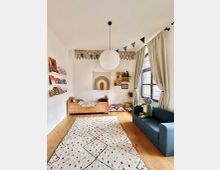 Kinderzimmer mit Holzfußboden, dekoriert mit einem weißen Teppich mit buntem Muster. Links steht ein Holzbett und Regale mit Büchern, rechts ein blaues Sofa vor einem großen Fenster mit beigen Vorhängen. Eine weiße Papierlampe hängt von der Decke, und Wimpelketten sowie kindgerechte Dekorationen schmücken den Raum.