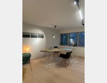 Minimalistisch eingerichteter Raum mit einem großen Tisch, der mit bunten Puzzleteilen bedeckt ist. Auf hellem Holzboden stehen moderne Stühle, eine Stehlampe, und an der weißen Wand hängen drei Schwarz-Weiß-Fotos; durch ein Fenster mit Pflanzen auf der Fensterbank ist eine bewaldete Außenansicht sichtbar.