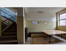 Ein einfacher Innenraum mit gelb-beigefarbenen Fliesenwänden, einem Holztisch mit Metallgestell und einer Bank an der Wand. Links führt eine Treppe mit rotem Geländer nach oben, und rechts befindet sich ein Fenster mit sichtbaren Außenziegelmauern.