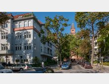 Straßenszene in einer urbanen Umgebung mit einem grauen Wohngebäude links, mehreren geparkten Autos und Bäumen, die den Gehweg säumen. Im Hintergrund ist eine Kirche mit einem spitzen, roten Turm zwischen den Bäumen sichtbar.