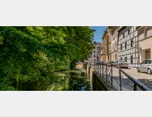 Schmaler, von Bäumen gesäumter Wasserlauf neben einer Kopfsteinpflasterstraße mit geparkten Autos und Fachwerk- sowie Altbauhäusern auf der rechten Seite. Ein schmiedeeisernes Geländer trennt den Gehweg vom Wasser.