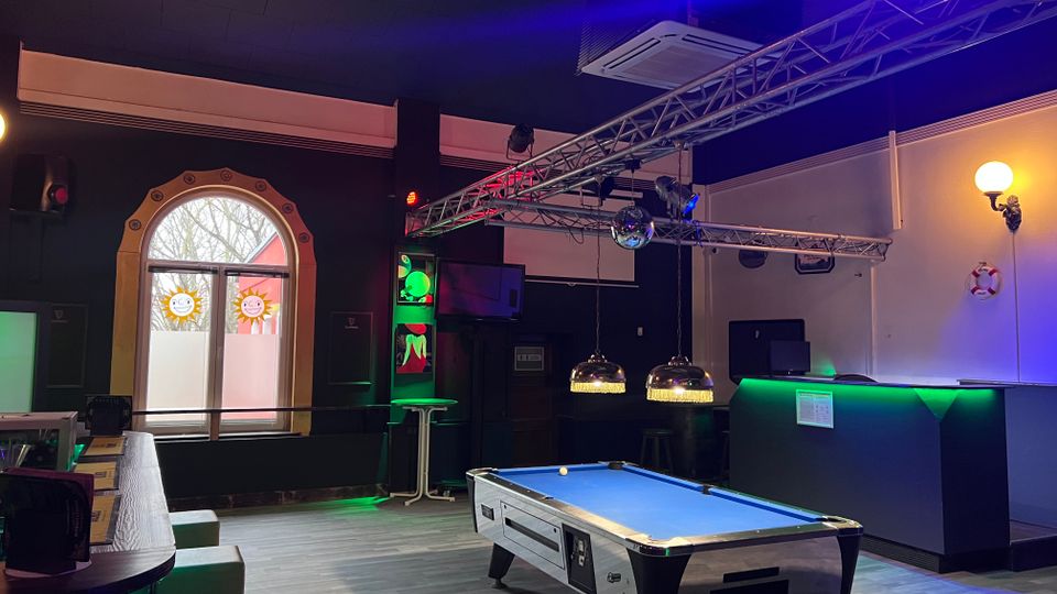 Innenraum einer Bar mit Billardtisch im Vordergrund, runden Tischen und Sitzbänken sowie einer Theke mit grünem Licht am rechten Rand. Über dem Raum hängen Traversen mit Bühnenbeleuchtung und Lampen; links ist ein großes Rundbogenfenster mit dekorierten Scheiben zu sehen.