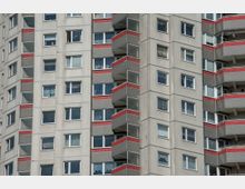 Hochhausfassade mit grauer Betonverkleidung, rechteckigen Fenstern und Balkonen, die durch rote und graue Paneele hervorgehoben sind. Die Balkone sind regelmäßig angeordnet und flankieren die Fensterflächen.