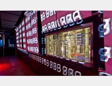 Ein dunkler Innenraum mit leuchtenden Projektionen von Zahlen und Symbolen auf den Wänden, einem Holzboden und einem Fenster, das den Blick in einen großen, industriellen Raum mit sichtbaren Säulen und einem Carlsberg-Logo freigibt.