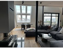 Modernes, offenes Wohnzimmer mit Essbereich unter einer Dachschräge, großen Fensterflächen und hellem Holzboden. Im Vordergrund stehen ein graues Ecksofa mit Marmorcouchtisch, daneben ein runder Esstisch; rechts führt eine Glastür auf einen Balkon.