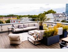 Moderne Dachterrasse mit Holzboden, grauen Loungemöbeln, einem kleinen Tisch mit Getränkekühler und dekorativen Pflanzen in Töpfen. Im Hintergrund sind Wohnhäuser, Grünanlagen und ein Hochhaus zu sehen.