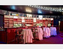 Eine stilvolle Bar mit roten Regalen, die mit Flaschen, Gläsern und Dekorationen bestückt sind. Vor der Bar stehen runde Tische mit weißen Tischdecken und hohen Barstühlen, darüber befinden sich Lichterketten.