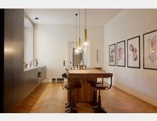 Moderne Küche mit minimalistischer Einrichtung, bestehend aus einer schwarzen Einbauküche auf der linken Seite und einem zentralen Holztisch mit Barhockern. An der rechten Wand hängen gerahmte, abstrakte Kunstdrucke, und drei hängende Pendelleuchten beleuchten den Raum.