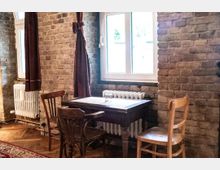 Kleiner Sitzbereich in einem Raum mit Ziegelsteinwänden, bestehend aus einem Holztisch und drei unterschiedlichen Stühlen. Im Hintergrund befinden sich ein Fenster mit weißen Rahmen, ein Heizkörper und dunkelrote Vorhänge.