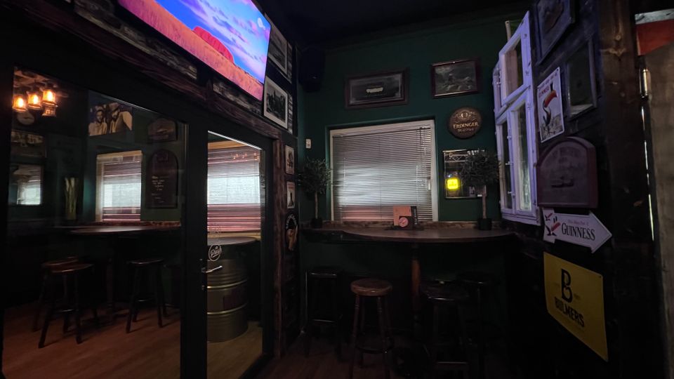 Gemütlicher Bar- oder Pub-Innenraum mit dunklen Holzvertäfelungen, hohen Barhockern und einem Wandfernseher über einem Spiegelbereich. An den Wänden hängen Bilder und Bierschilder; ein Fenster mit Jalousie sowie ein geöffnetes Fenster mit Läden prägen die Rückwand.