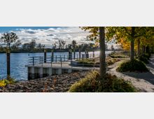 Uferpromenade mit gepflastertem Weg und Bäumen, die teilweise herbstlich gefärbt sind. Im Hintergrund befindet sich ein Steg mit Geländer, der ins Wasser eines ruhigen Flusses ragt, sowie einzelne Bojen und eine bewaldete Flusslandschaft.