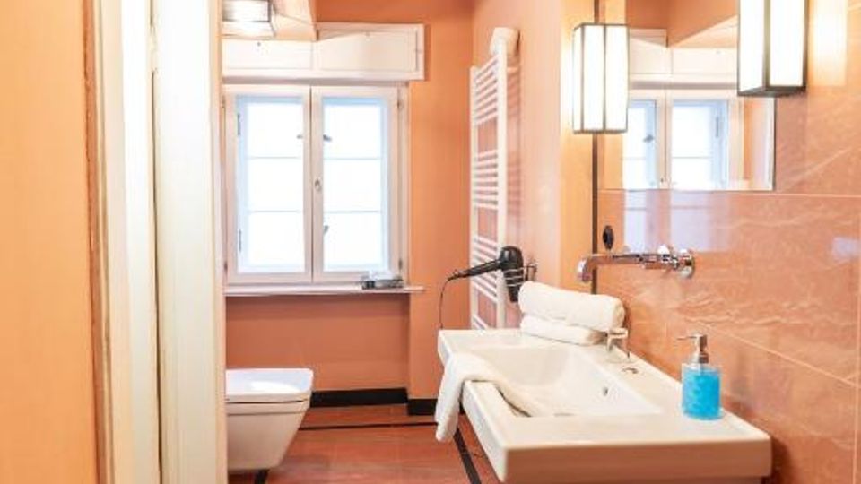 Langes Badezimmer mit rosafarbenen Fliesen, schwarzer Bodenbordüre und einem weißen Waschtisch mit großem Wandspiegel und rechteckigen Wandleuchten. Am Ende des Raums stehen ein WC und ein Fenster, daneben ein Handtuchheizkörper.