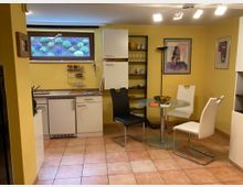 Kleine Küche mit gelb gestrichenen Wänden, weißen Einbauschränken, einem kleinen Fenster mit Blick auf einen bepflanzten Außenbereich und Fliesenboden. Im Vordergrund ein runder Glastisch mit drei Stühlen, daneben ein Regal mit Geschirr und Dekorationsgegenständen.