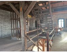 Ein Raum mit rustikalem Interieur, geprägt von Holzbalken und einer offenen Holztreppe, die zu einer oberen Ebene führt. Die Wände sind aus unbehandeltem Stein, und an der Seite steht eine Reihe von gestapelten Holzstühlen vor großen Fenstern mit abgerundeten Bögen, die Tageslicht hereinlassen.