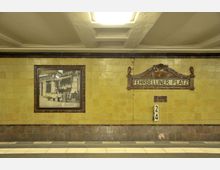 U-Bahn-Station mit gelb gekachelter Wand, einem historischen Schild mit der Aufschrift "Fehrbelliner Platz" und einer alten Schwarz-Weiß-Fotografie in einem Rahmen. Der Boden besteht aus grauen Fliesen mit weißen Streifen.