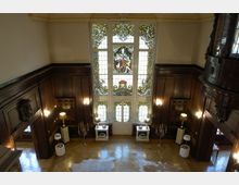 Großer Innenraum mit hölzernen Wandvertäfelungen und einem kunstvoll gestalteten Buntglasfenster mit Szenen und Ornamenten. Der Boden ist aus poliertem Holz, und der Raum ist mit Stehlampen, Marmorsockeln und Kerzenständern dekoriert.