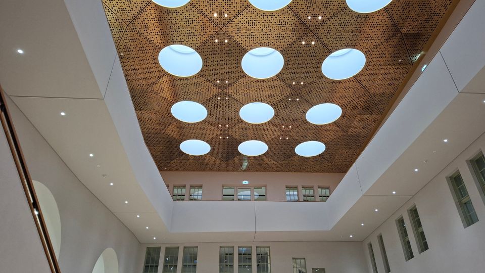 Innenansicht eines modernen Atriums mit hoher Decke aus perforierten Holzpaneelen und mehreren runden Oberlichtern. Umgeben von hellen Wänden mit integrierten Spotlights sowie Fensterreihen und Galerien auf mehreren Ebenen.