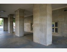 Überdachter Bereich mit quadratischen, geriffelten Steinsäulen und einer Wand aus hellen Steinfliesen mit vergitterten Fenstern. Im Hintergrund sind Teile eines Außenbereichs und Bäume sichtbar.