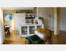 Wohnbereich mit einer offenen, modernen Küche in Weiß, einer Kochinsel mit Milchglastüren, und einer Essgruppe links im Hintergrund. Im Vordergrund steht ein brauner Ledersessel mit einer grünen Decke, der Raum hat einen hellen Parkettboden und ist mit Dekor wie Pflanzen und einem Küstenbild an der Wand gestaltet.