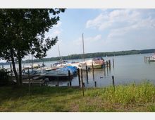 Bootsanleger an einem See mit mehreren festgemachten kleinen Segel- und Motorbooten, teils mit Abdeckplanen. Im Vordergrund befindet sich eine grasbewachsene Fläche, im Hintergrund eine Waldlinie und ein teils bewölkter Himmel.