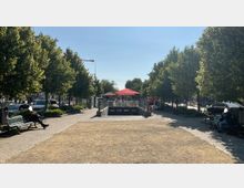Städtischer Platz mit einer breiten, von Bäumen gesäumten Fußgängerzone und einem Eingang zu einer U-Bahn-Station in der Mitte. Im Hintergrund sind Sonnenschirme vor einem Cafébereich sichtbar, und Autos parken entlang der Straße.