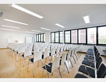 Moderner Seminarraum mit mehreren Reihen weißer Stühle auf hellem Holzboden, großen Fenstern an der rechten Seite, durch die Gebäude und Bäume sichtbar sind. Im Vordergrund steht ein Rednerpult sowie Flipcharts an der weißen Wand.