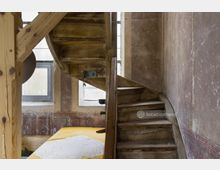 Holztreppe in einem historischen Gebäude mit abgenutzten Stufen und grober Wandstruktur. Im Hintergrund sind kleine Fenster mit einfacher Verglasung sowie ein gelber Tisch mit einer Tischdecke sichtbar.