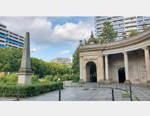 Ein Außenbereich mit einem steinernen Obelisken links und einem historischen Pavillon mit Bögen und Säulen rechts. Im Hintergrund befinden sich moderne Wohn- und Bürogebäude sowie Bäume und ein parkähnliches Umfeld.