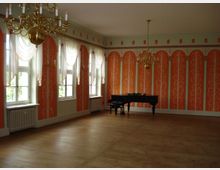 Ein prunkvoll gestalteter Saal mit orangefarbenen Wänden, die mit weißen Ornamenten verziert sind. Im Raum stehen ein schwarzer Flügel und mehrere Kronleuchter aus Gold, während durch hohe Fenster mit weißen Vorhängen Tageslicht einfällt.