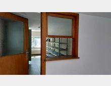 Ein heller Innenraum mit einer offenen Holztür und einem Wanddurchbruch mit Glaseinsatz. Im Hintergrund befinden sich Metallregale, ein Heizkörper unter einem Fenster und ein heller Bodenbelag.