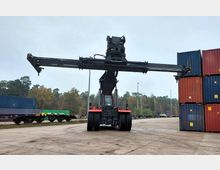 Ein großer Containerstapler hebt einen Container in einem Güterterminal, umgeben von gestapelten blauen und roten Containern sowie Bahnwaggons. Im Hintergrund sind Bäume und ein bewölkter Himmel zu sehen.