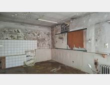 Verlassener Innenraum mit stark abgeplatzter Farbe und feuchten, fleckigen Wänden, teils mit weißen Wandfliesen und einem beschädigten Waschbecken. An der Decke hängt eine Leuchtstoffröhre; an der Wand sind ein leerer Schaukasten sowie ein alter Heizkörper und ein kleines Regal zu sehen.
