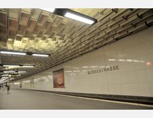 U-Bahn-Station mit dem Schriftzug „Blissestraße“ an einer weißen, gefliesten Wand. Die Decke ist mit einem wabenartigen Muster gestaltet, und moderne Leuchtstofflampen beleuchten den Bahnsteig.