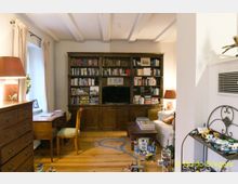 Wohnzimmer mit Holzmöbeln und Bücherschrank. Im Raum befinden sich ein Schreibtisch mit Stuhl, eine Couch, ein Couchtisch, Spielzeug auf dem Holzboden und ein Bücherregal mit integriertem Fernseher.