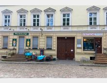 Fassade eines historischen Gebäudes mit gelber Putzfassade und sechs Fenstern im Obergeschoss. Im Erdgeschoss befinden sich links die "Pferdekneipe" mit blau eingedeckten Tischen und rechts ein Laden mit dem Schild "Fritsche Stickerei & Druck", dazwischen eine große Holztür.