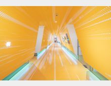 Ein futuristisch gestalteter Innenkorridor mit leuchtend gelben Wänden, die mit weißen Punkt-Mustern versehen sind. Die Struktur hat schräge Seitenwände, einen glänzenden Boden und integrierte LED-Beleuchtung entlang der unteren Kanten.