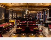 Elegantes Innenraumdesign eines Restaurants oder einer Bar mit dunkler Holzausstattung, roten Polstersitzbänken, kleinen Tischen mit Kerzen und einem goldenen Disco-Ball an der Decke. Warme Beleuchtung und große Fenster schaffen eine gemütliche Atmosphäre.
